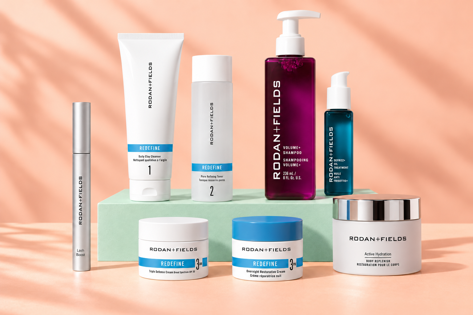 Rodan + Fields REDEFINE regimen, Volume+ Shampoo, Defrizz+ Oil Treatment, Lash Boost, and Body Replenish on a peach tabletop and mint riser